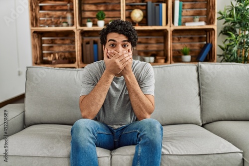 Young hispanic man wearing casual clothes sitting on the sofa at home shocked covering mouth with hands for mistake. secret concept. © Krakenimages.com