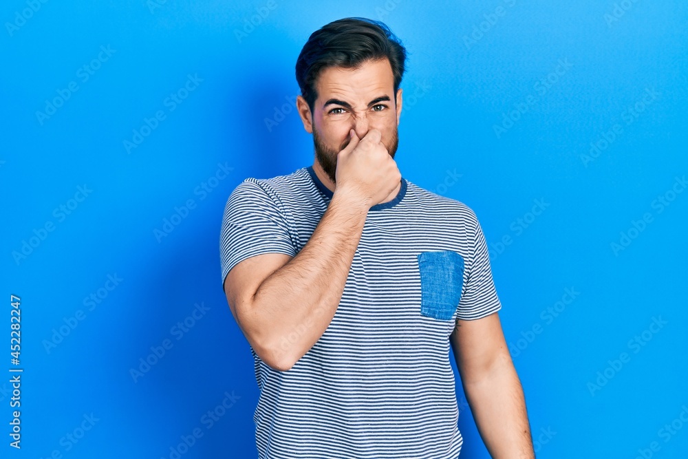 Handsome caucasian man with beard wearing casual striped t shirt smelling something stinky and disgusting, intolerable smell, holding breath with fingers on nose. bad smell Handsome caucasian man with beard wearing casual striped t shirt smelling something stinky and disgusting, intolerable smell, holding breath with fingers on nose. bad smell