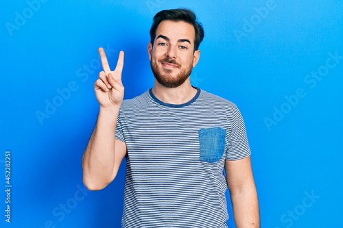 Handsome caucasian man with beard wearing casual striped t shirt showing and pointing up with fingers number two while smiling confident and happy.