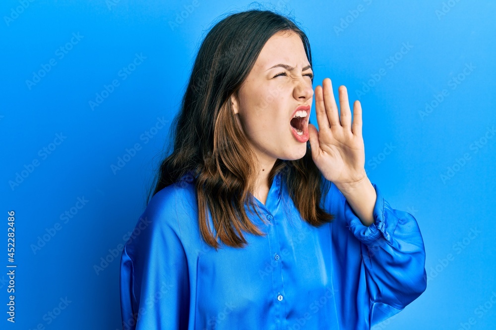 Young brunette woman wearing casual blue shirt shouting and screaming loud to side with hand on mouth. communication concept. Young brunette woman wearing casual blue shirt shouting and screaming loud to side with hand on mouth. communication concept.