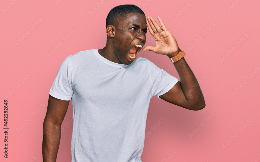 Young african american man wearing casual white t shirt shouting and screaming loud to side with hand on mouth. communication concept. Young african american man wearing casual white t shirt shouting and screaming loud to side with hand on mouth. communication concept.