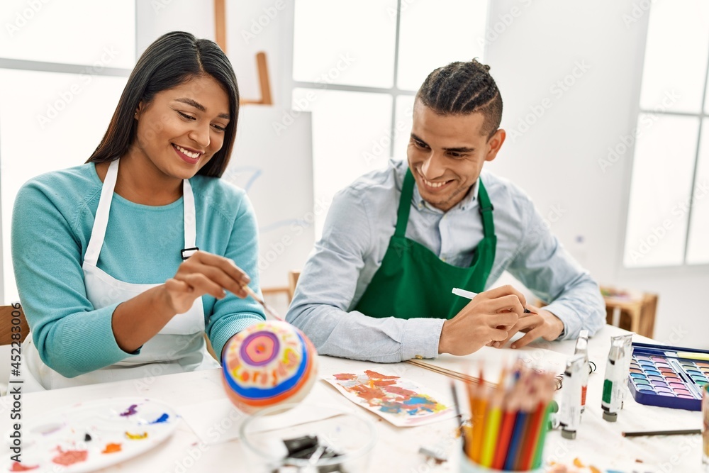 Fototapeta premium Young latin painter couple smiling happy painting sitting on the table at art studio