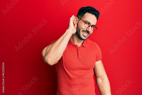 Young hispanic man wearing casual clothes and glasses smiling with hand over ear listening an hearing to rumor or gossip. deafness concept.