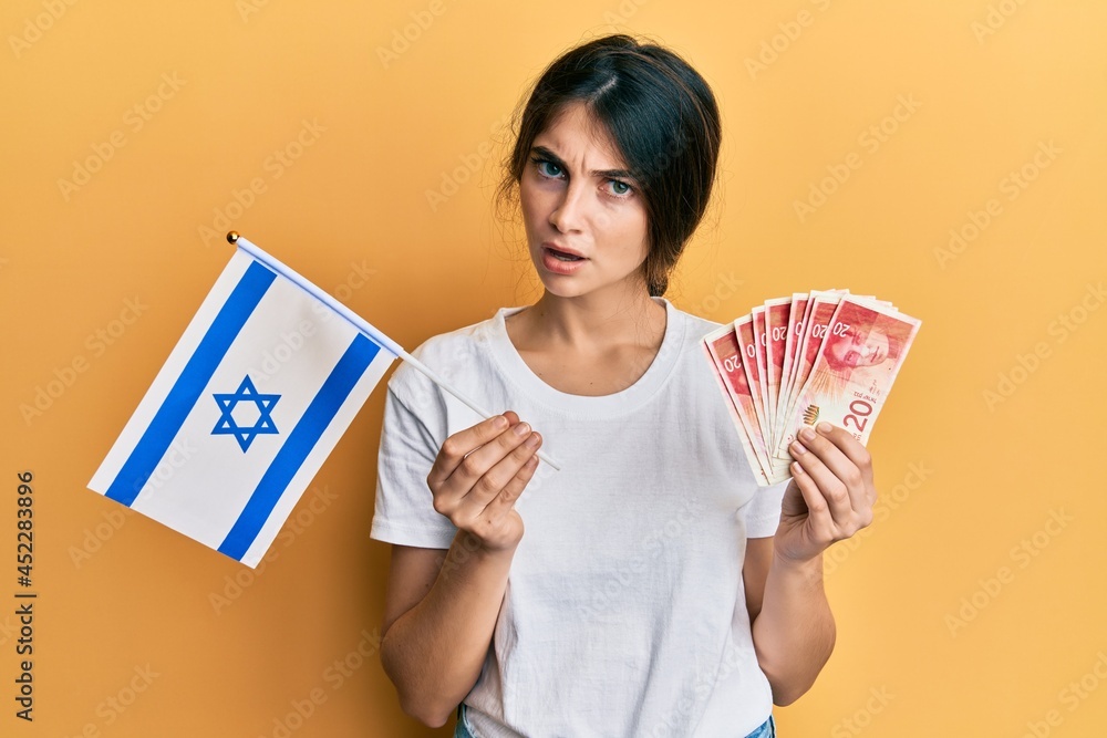 Young caucasian woman holding israel flag and shekels banknotes in shock face, looking skeptical and sarcastic, surprised with open mouth Young caucasian woman holding israel flag and shekels banknotes in shock face, looking skeptical and sarcastic, surprised with open mouth