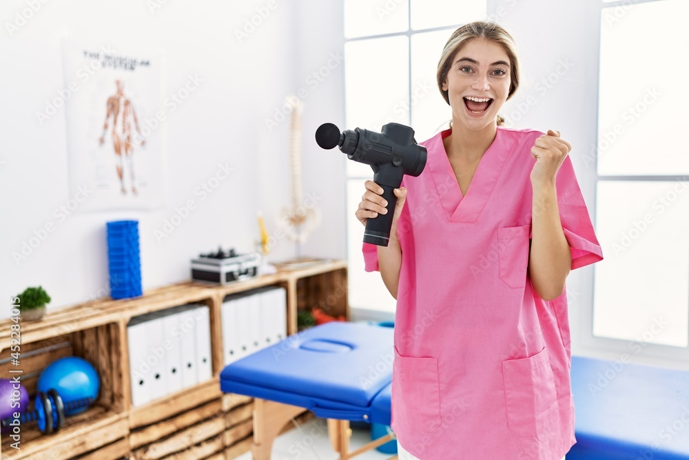 Young physiotherapist woman holding therapy massage gun at wellness center screaming proud, celebrating victory and success very excited with raised arms