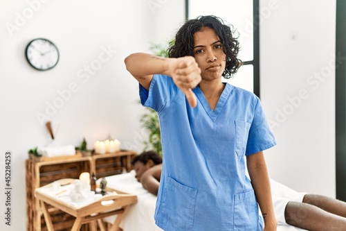 Young therapist woman at wellness spa center looking unhappy and angry showing rejection and negative with thumbs down gesture. bad expression.