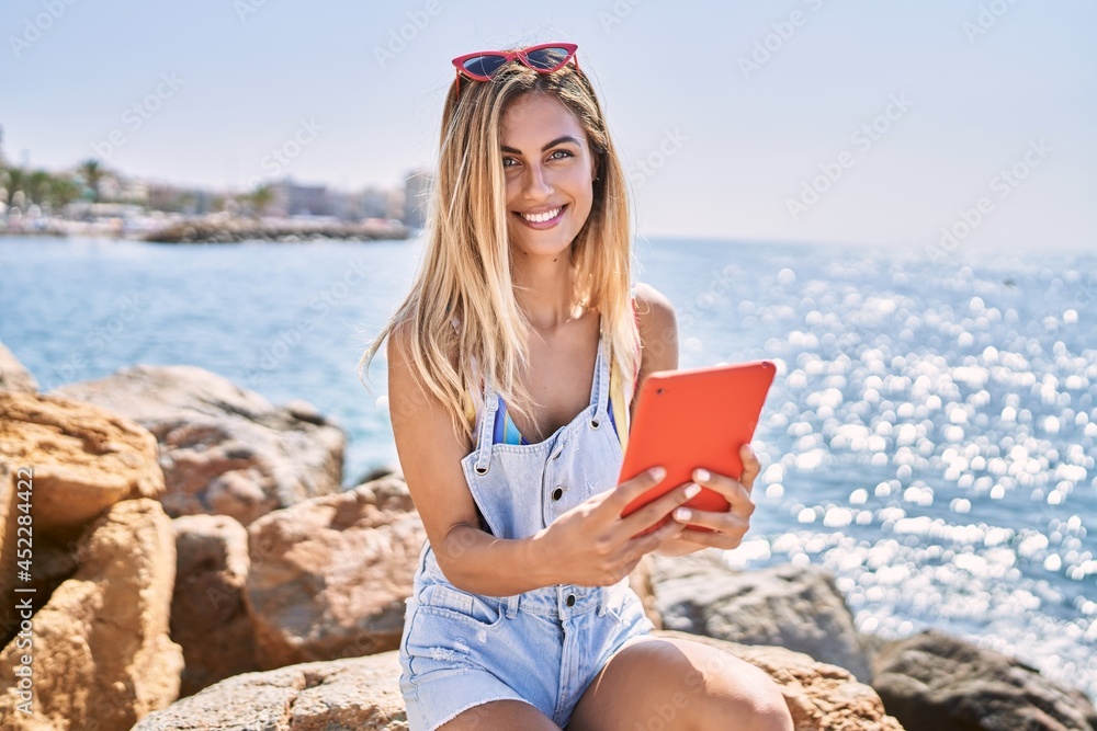 Young blonde girl using touchpad sitting on the rock at the beach.