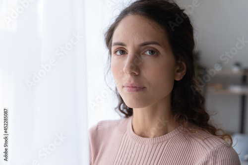 Close up portrait of joyless 30s woman standing alone near window indoor. Face of Hispanic serious female with sad eyes staring at camera. Tiredness, lack of optimism, solitude, life concerns concept photo