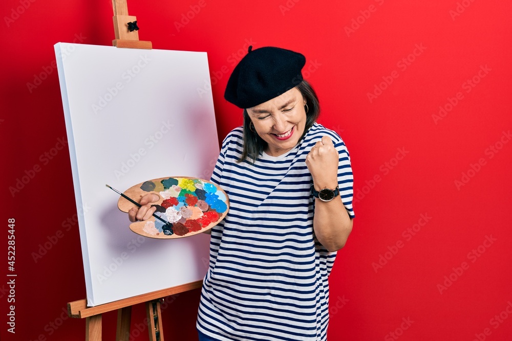 Fototapeta premium Middle age hispanic woman standing drawing with palette by painter easel stand very happy and excited doing winner gesture with arms raised, smiling and screaming for success. celebration concept.