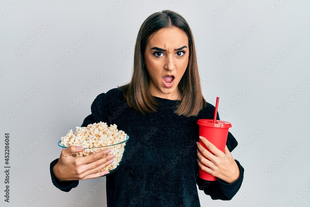 Young brunette girl eating popcorn and drinking soda in shock face, looking skeptical and sarcastic, surprised with open mouth