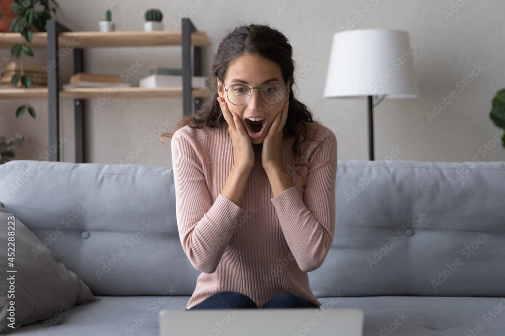 Young excited woman in eyeglasses open mouth while read unbelievable incredible media news. Amazed shocked surprised lady staring at laptop screen cant believe in lottery luck gambling victory concept