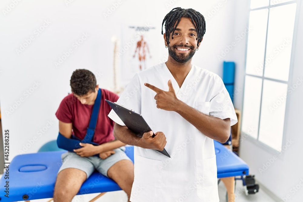 Obraz premium Young hispanic man working at pain recovery clinic with a man with broken arm cheerful with a smile on face pointing with hand and finger up to the side with happy and natural expression