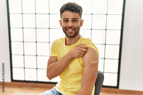 Arab young man getting vaccine showing arm with band aid looking positive and happy standing and smiling with a confident smile showing teeth photo