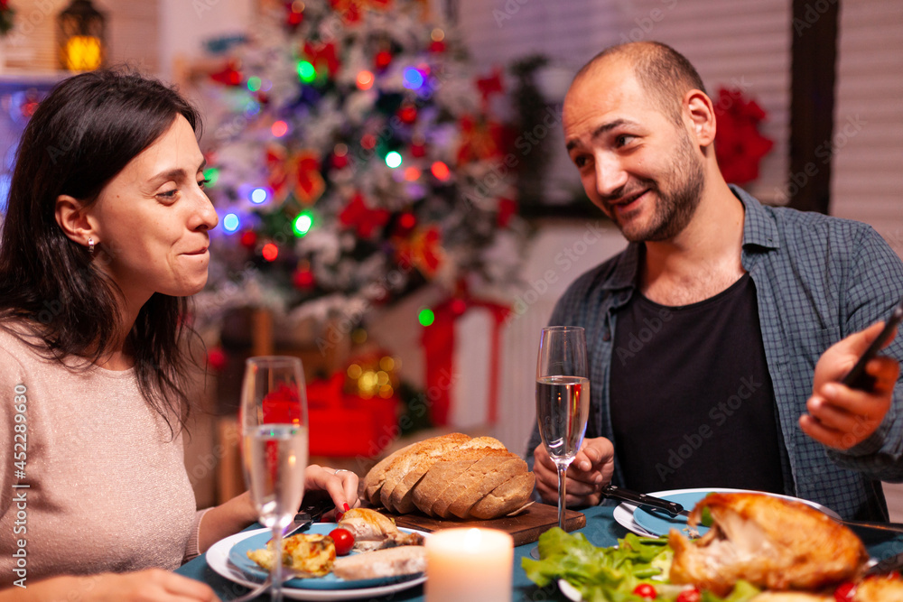 Happy family eating delicious dinner sitting at dining table in xmas decorated kitchen celebrating christmas holiday. Couple watching entertainment movie show using phone. Santa-claus season Happy family eating delicious dinner sitting at dining table in xmas decorated kitchen celebrating christmas holiday. Couple watching entertainment movie show using phone. Santa-claus season