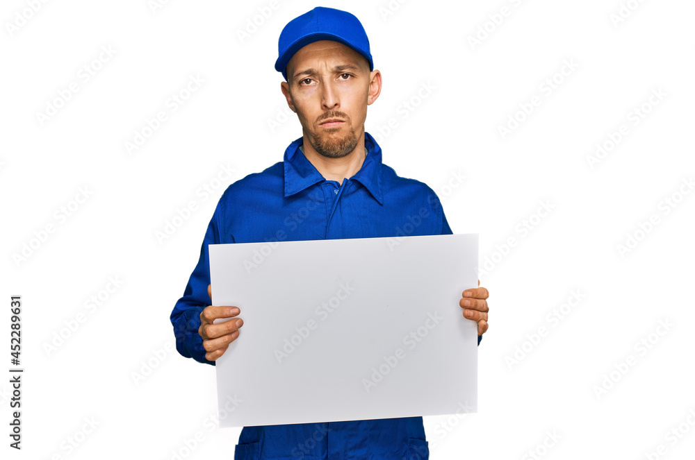 Bald man with beard wearing builder jumpsuit uniform holding empty banner thinking attitude and sober expression looking self confident Bald man with beard wearing builder jumpsuit uniform holding empty banner thinking attitude and sober expression looking self confident