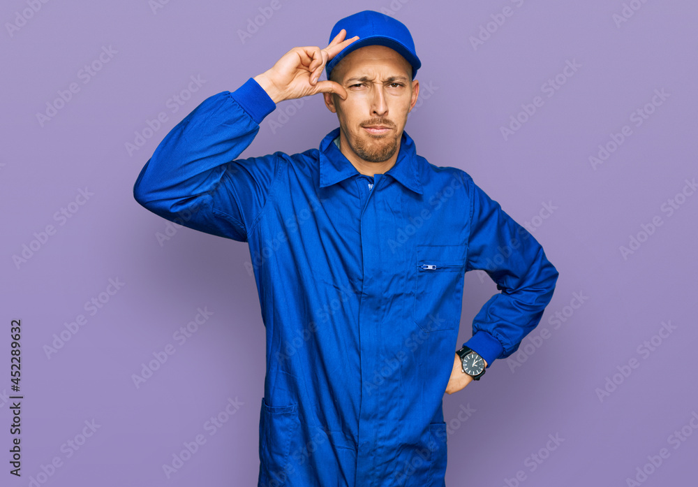 Bald man with beard wearing builder jumpsuit uniform worried and stressed about a problem with hand on forehead, nervous and anxious for crisis