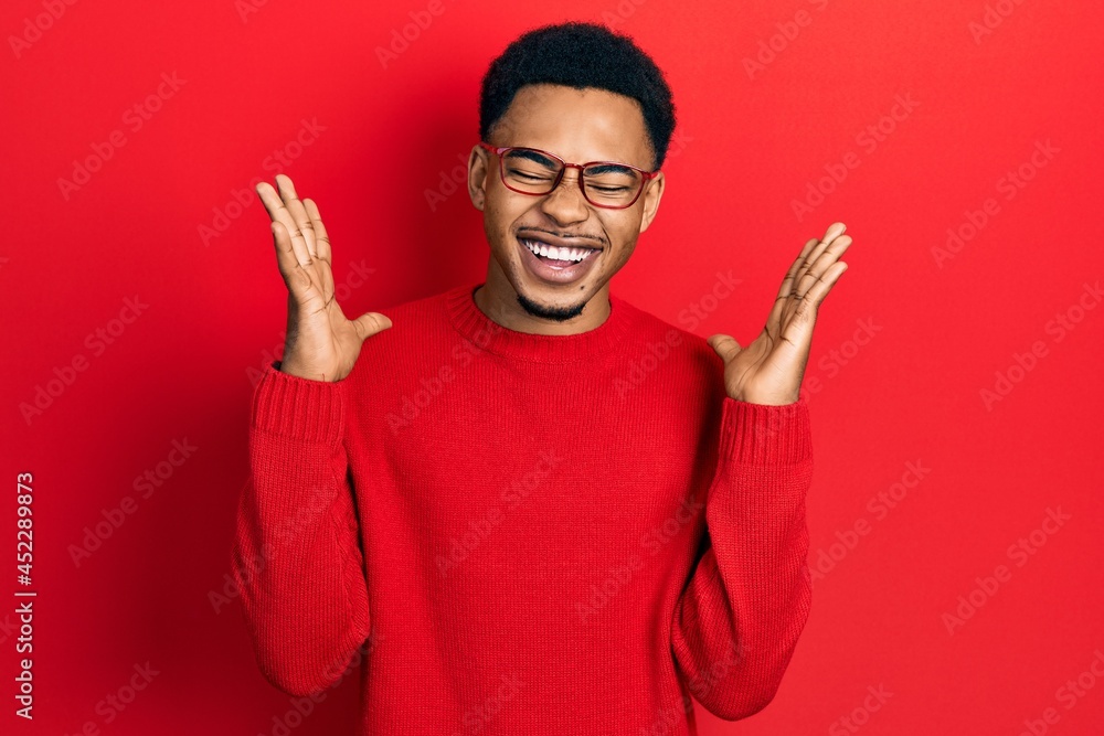 Naklejka premium Young african american man wearing casual clothes and glasses celebrating mad and crazy for success with arms raised and closed eyes screaming excited. winner concept