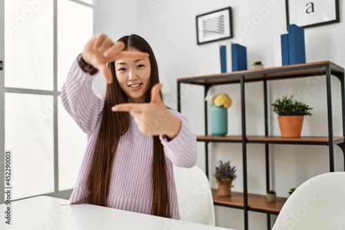 Young chinese girl wearing casual clothes sitting on the table at home smiling making frame with hands and fingers with happy face. creativity and photography concept.