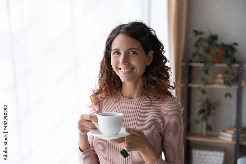 Happy young Latina woman standing in cozy room smile looks at camera holds cup of fresh brewed tea, enjoy no stress calm positive pastime, new day drink favourite beverage. Leisure, lifestyle concept Happy young Latina woman standing in cozy room smile looks at camera holds cup of fresh brewed tea, enjoy no stress calm positive pastime, new day drink favourite beverage. Leisure, lifestyle concept
