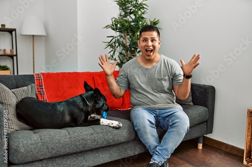Young latin man and dog sitting on the sofa at home celebrating crazy and amazed for success with arms raised and open eyes screaming excited. winner concept © Krakenimages.com