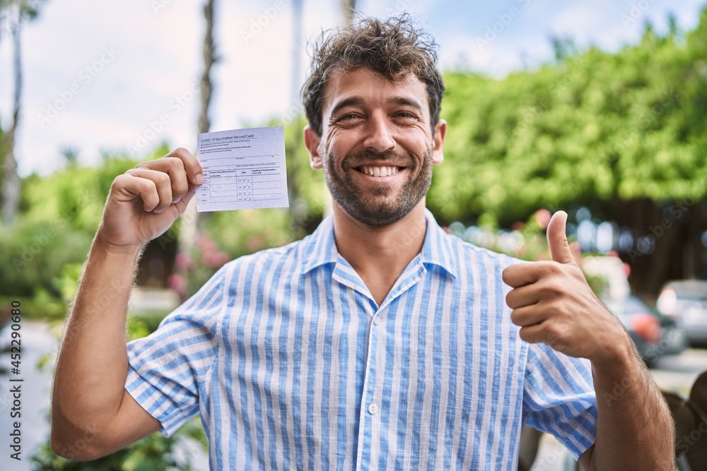 Fototapeta premium Young handsome man holding covid record card smiling happy and positive, thumb up doing excellent and approval sign