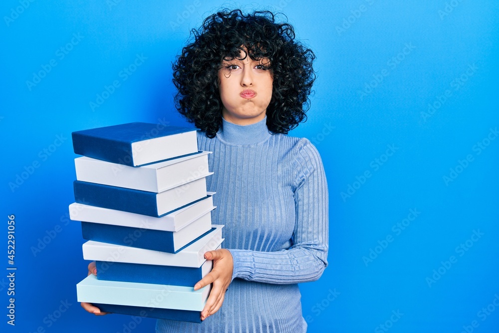 Young middle east woman holding a pile of books puffing cheeks with funny face. mouth inflated with air, catching air. Young middle east woman holding a pile of books puffing cheeks with funny face. mouth inflated with air, catching air.