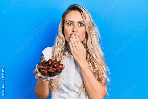 Beautiful young blonde woman holding bowl with dates covering mouth with hand  shocked and afraid for mistake. surprised expression