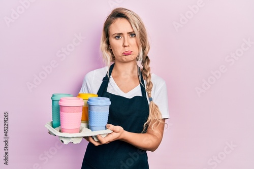 Beautiful young blonde woman wearing waitress apron holding take away cup of coffees depressed and worry for distress, crying angry and afraid. sad expression.