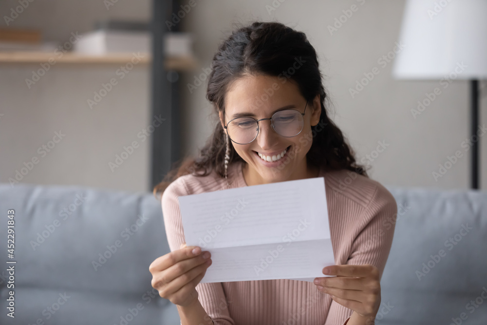 Smiling attractive young woman in glasses sit on sofa at home reading letter. Pleased female holds in hands sheet pleased by good news written in official document. Bank notice, happy client concept Smiling attractive young woman in glasses sit on sofa at home reading letter. Pleased female holds in hands sheet pleased by good news written in official document. Bank notice, happy client concept
