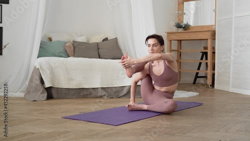 Slender, flexible, caucasian middle-aged woman with black hair in beige tracksuit performs standing asanas one-legged king pigeon pose on purple yoga mat in bright room with bed, a mirror, a window photo