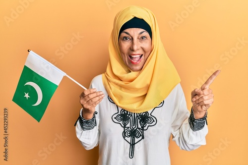 Middle age hispanic woman wearing hijab holding pakistan flag smiling happy pointing with hand and finger to the side