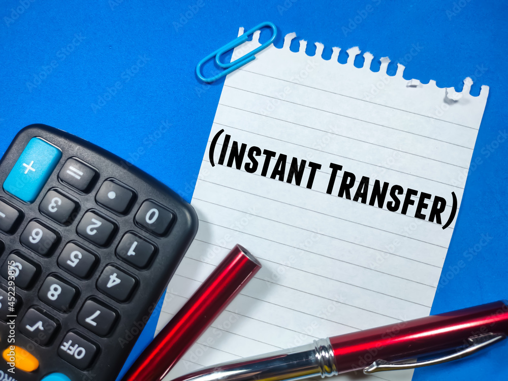Business concept.Text INSTANT TRANSFER writing on notepaper with open pen and calculator on blue background. Business concept.Text INSTANT TRANSFER writing on notepaper with open pen and calculator on blue background.