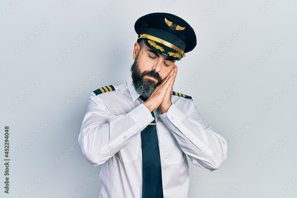 Young hispanic man wearing airplane pilot uniform sleeping tired dreaming and posing with hands together while smiling with closed eyes. Young hispanic man wearing airplane pilot uniform sleeping tired dreaming and posing with hands together while smiling with closed eyes.