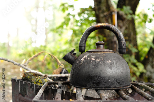 Old metal tea pot covered with soot and dust on grill. Making tea in nature camping in the forest. Cooking on fire in summer.