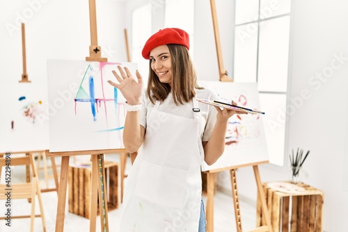 Young artist woman painting on a canvas at art studio waiving saying hello happy and smiling, friendly welcome gesture