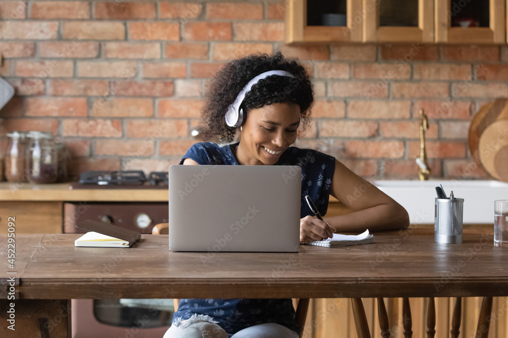 Happy Black student girl in wireless headphones writing notes, watching ...