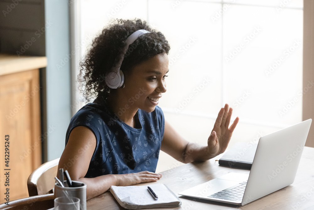 Φωτογραφία Happy African American remote teacher, coach in wireless headphones waving at laptop webcam, greeting audience, students, giving virtual training, online seminar, webinar