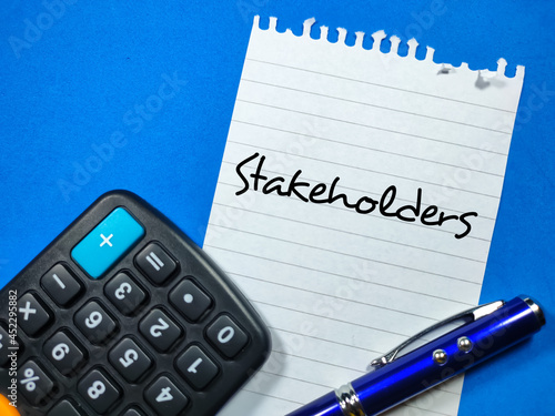 Business concept.Text Stakeholders writing on notepaper with pen and calculator on blue background.