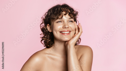Smiling sensual woman with curly natural hair, glowing face and healhy skin, shiny body after shower. Happy girl touches her smooth and gentle face after using skin care product, pink background