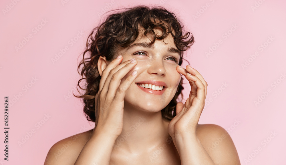 Beautiful natural girl smiling, rubbing her face with facial cleanser for glowing healthy skin, looking happy. Curly young woman showering, using skincare hydrating products, pink background - obrazy, fototapety, plakaty 