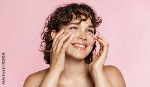 Beautiful natural girl smiling, rubbing her face with facial cleanser for glowing healthy skin, looking happy. Curly young woman showering, using skincare hydrating products, pink background photo