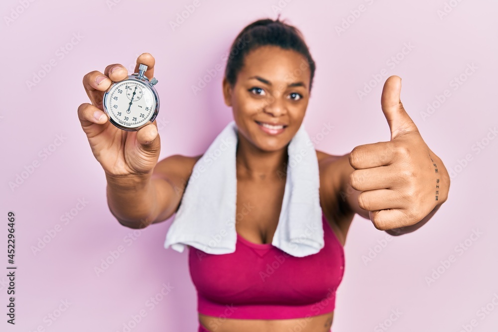 Young african american girl wearing sportswear holding stopwatch smiling happy and positive, thumb up doing excellent and approval sign Young african american girl wearing sportswear holding stopwatch smiling happy and positive, thumb up doing excellent and approval sign