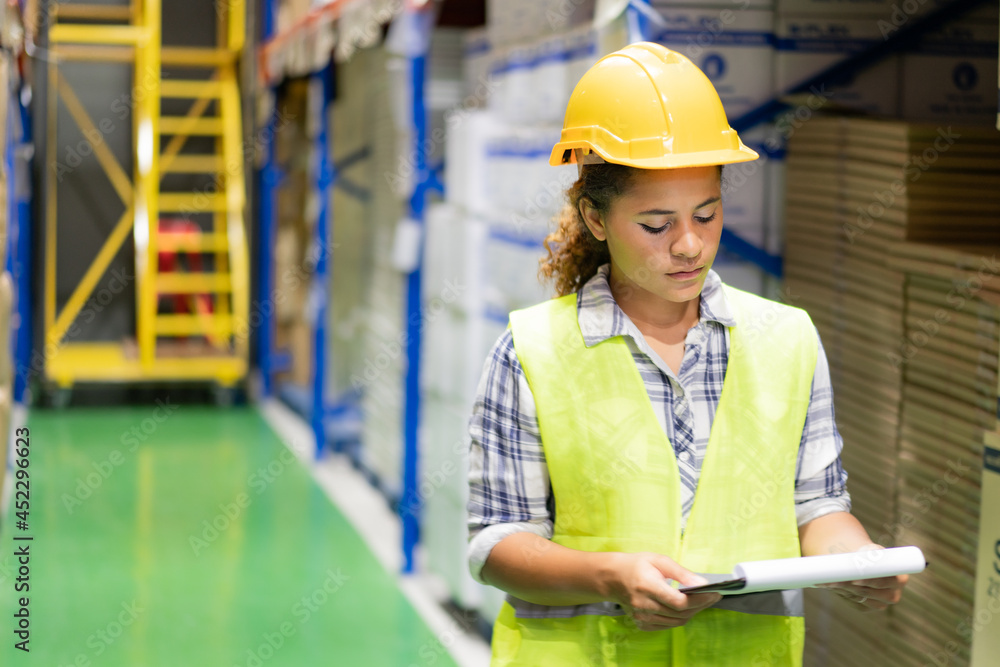 Young pretty African female warehouse worker in a yellow safety vest and helmet standing at a warehouse aisle looking at a clipboard in hand while doing stocktaking of products in a blurred warehouse. Young pretty African female warehouse worker in a yellow safety vest and helmet standing at a warehouse aisle looking at a clipboard in hand while doing stocktaking of products in a blurred warehouse.