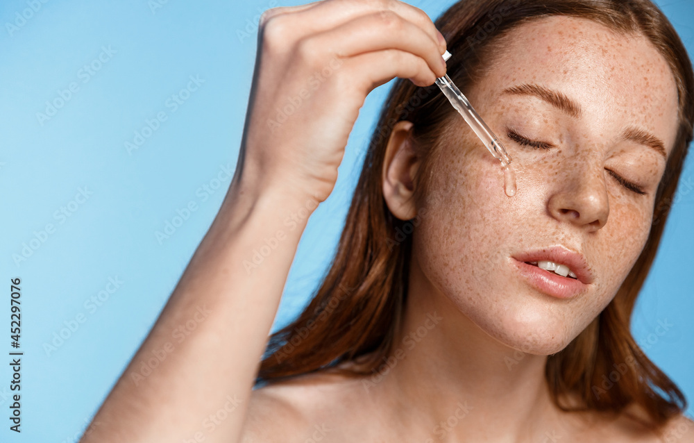 Close up of redhead girl applying facial serum with dropper, cleansing and moisturizing lotion for daily skin care routine, standing over blue background