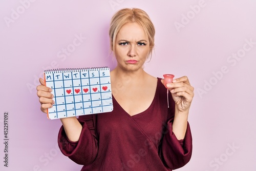 Young blonde woman holding heart calendar and menstrual cup depressed and worry for distress, crying angry and afraid. sad expression.