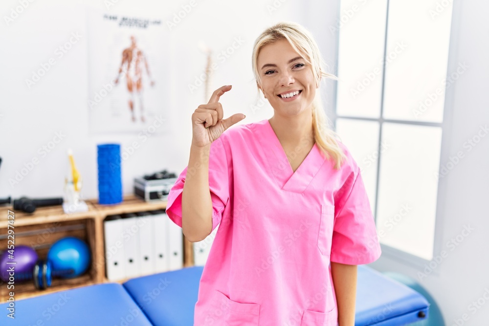 Young caucasian woman working at pain recovery clinic smiling and confident gesturing with hand doing small size sign with fingers looking and the camera. measure concept. Young caucasian woman working at pain recovery clinic smiling and confident gesturing with hand doing small size sign with fingers looking and the camera. measure concept.