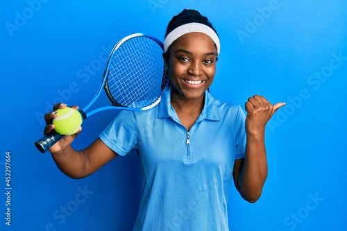 African american woman with braided hair playing tennis holding racket and ball pointing thumb up to the side smiling happy with open mouth © Krakenimages.com