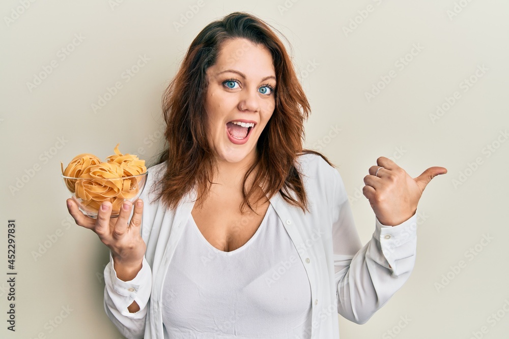 Young caucasian woman holding bowl of uncooked pasta pointing thumb up to the side smiling happy with open mouth