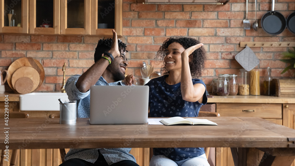 © fizkes - Happy successful Afro American couple celebrating financial achieve at laptop, giving high five. Married man and woman getting income, profit, loan, mortgage bank approval, feeling joy together