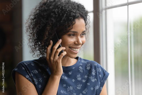 Happy dreamy young African American woman talking on mobile phone, looking out of window, smiling at thoughts, getting, discussing good news, thinking over telephone call, enjoying conversation